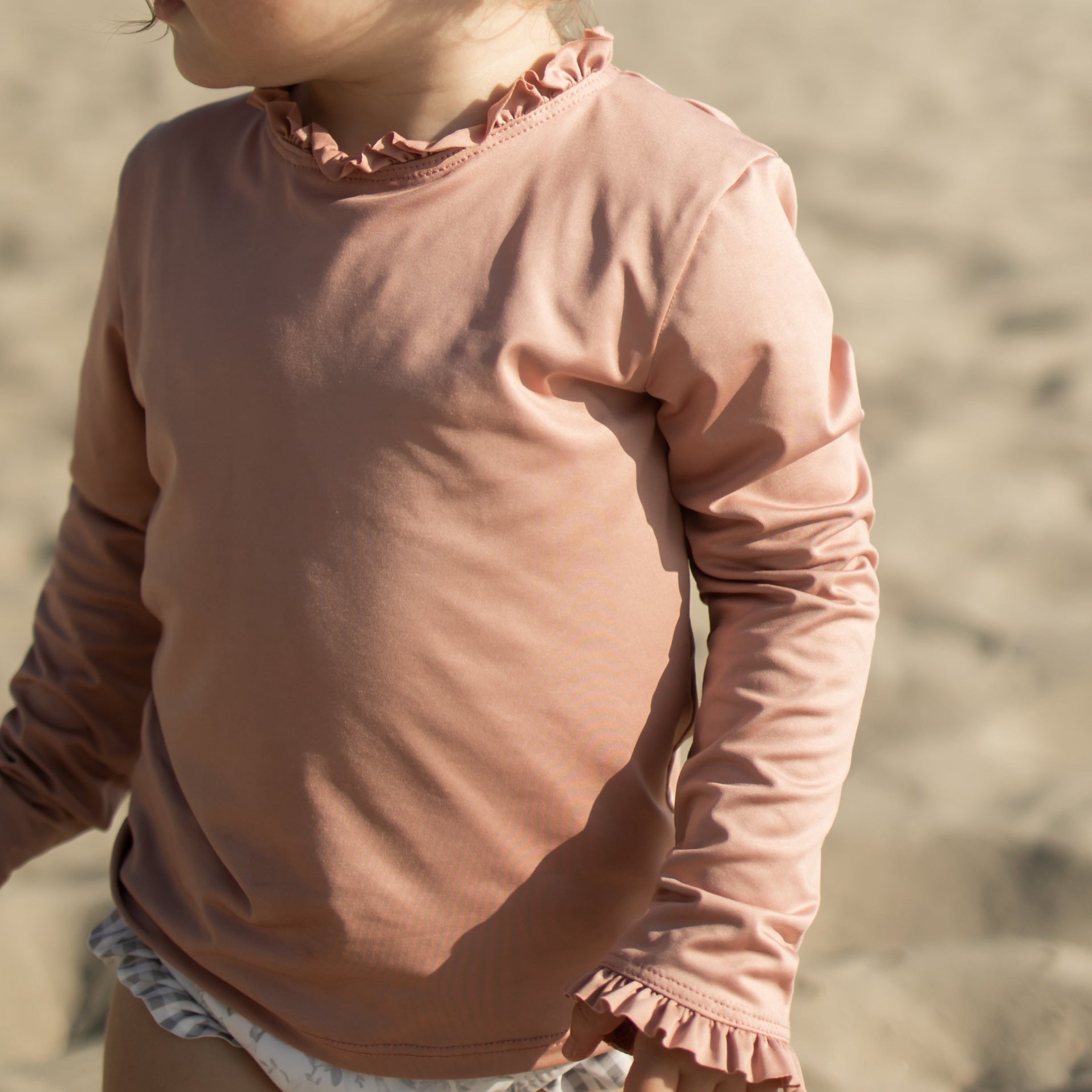 Child wearing a pink long-sleeve top and white shorts on a sandy background