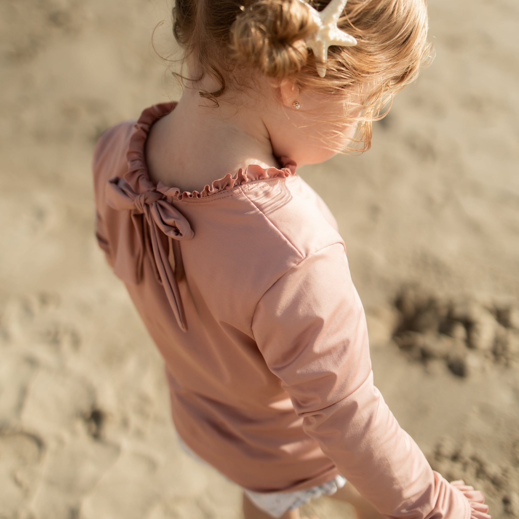 Child in a pink jacket standing on a sandy beach with a starfish in their hair.