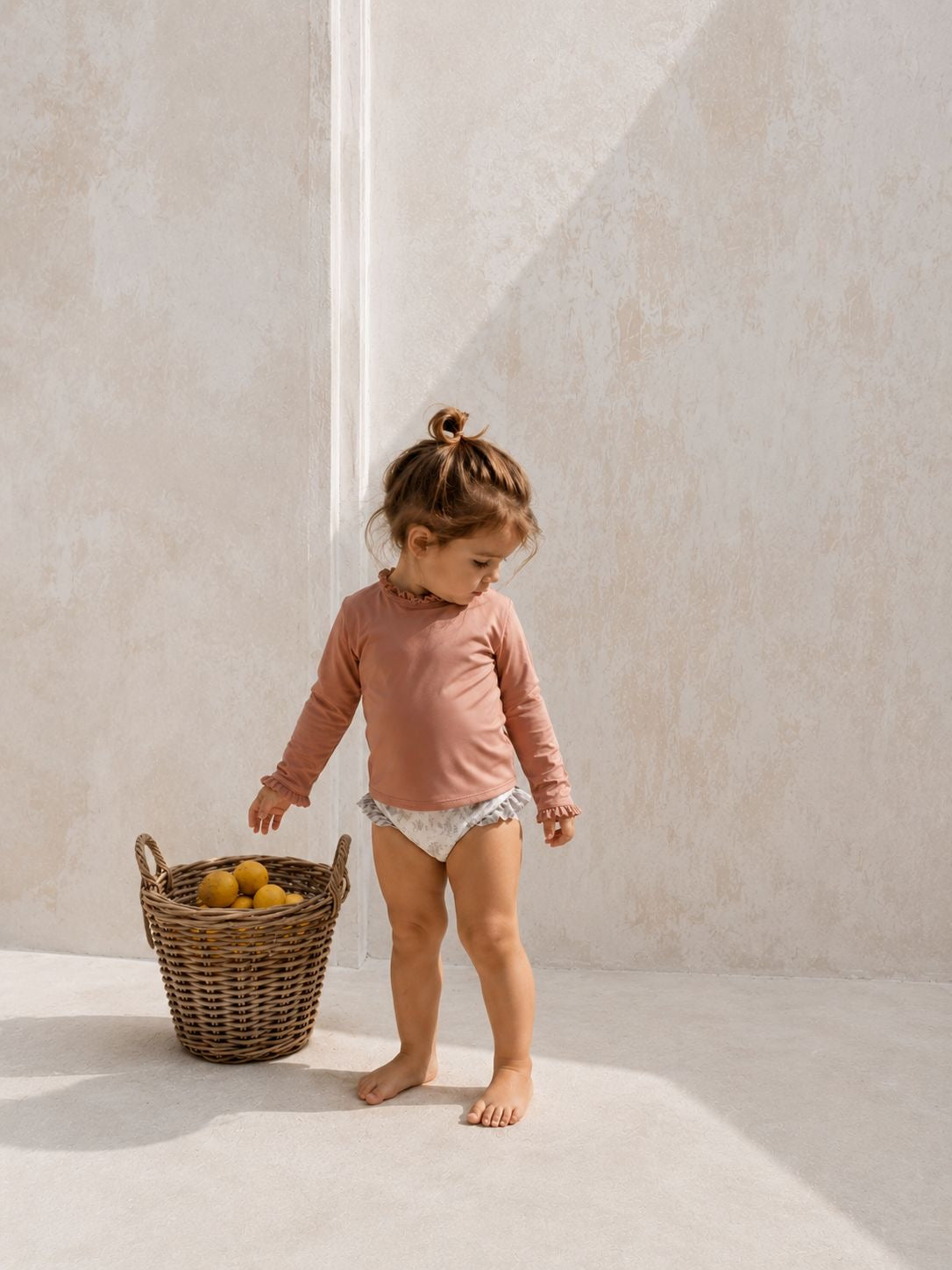 Child in a pink shirt standing next to a wicker basket with fruits on a light background