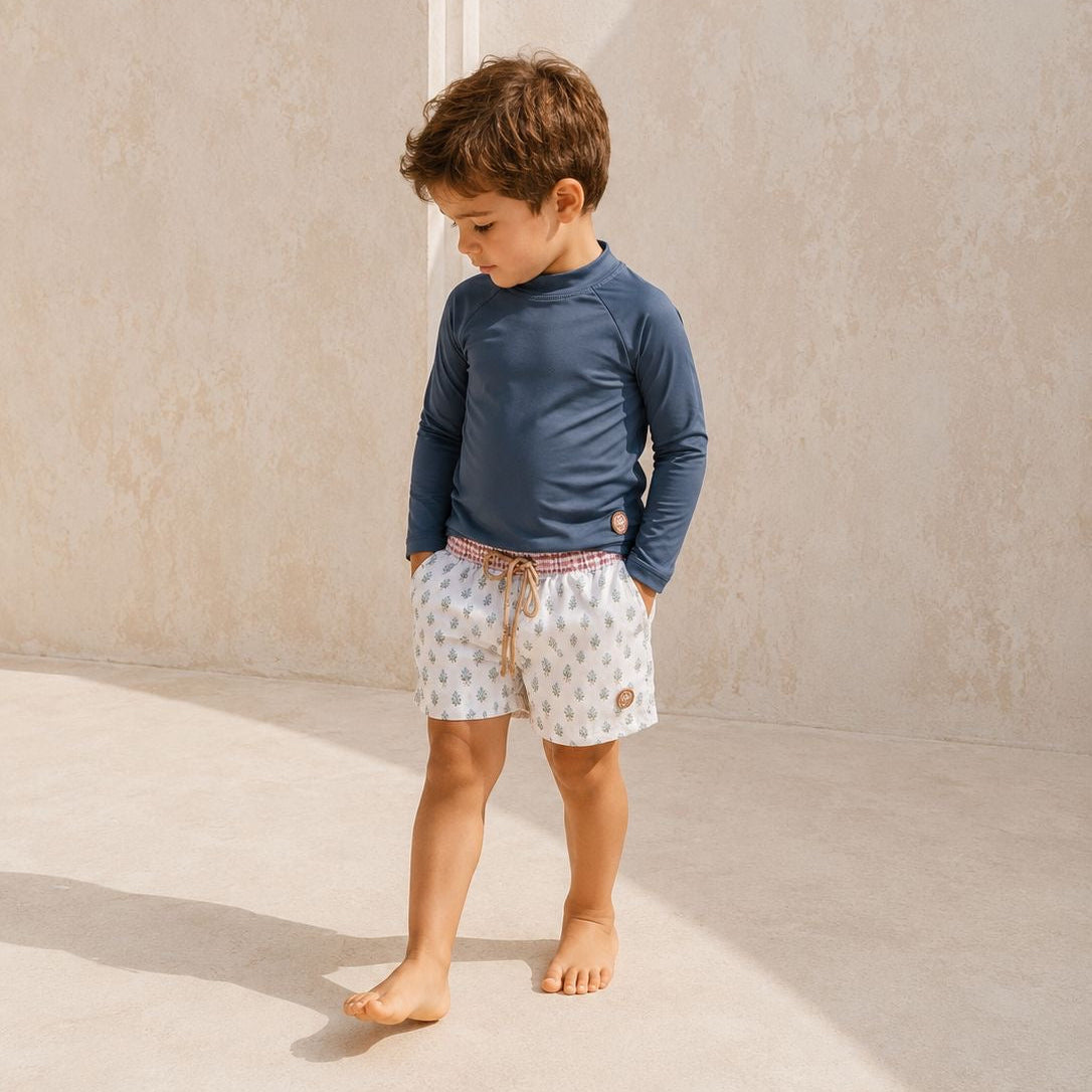 Young boy wearing a blue long-sleeve shirt and patterned shorts standing against a beige wall.